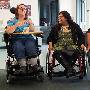 Two women in wheelchairs and a woman with a service dog sit together in a classroom