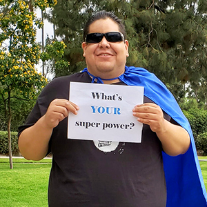 A man wearing a cape holds a sign that says, What's YOUR superpower?