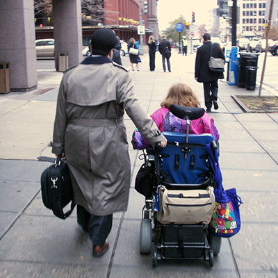 The backs of two people on a sidewalk, one of them walking and one of them in a power wheelchair