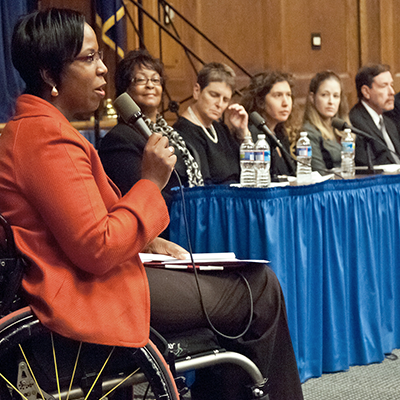 Woman in a wheelchair speaks into a microphone with an audience in the background